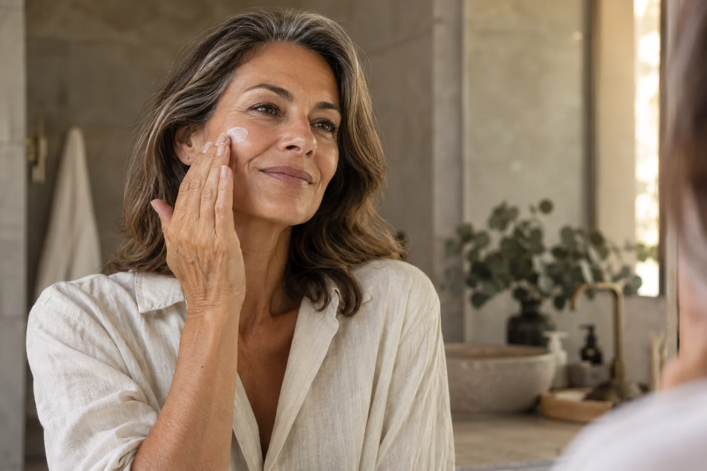 Vrouw van middelbare leeftijd die moisturizer op haar wang aanbrengt in een rustige badkamer